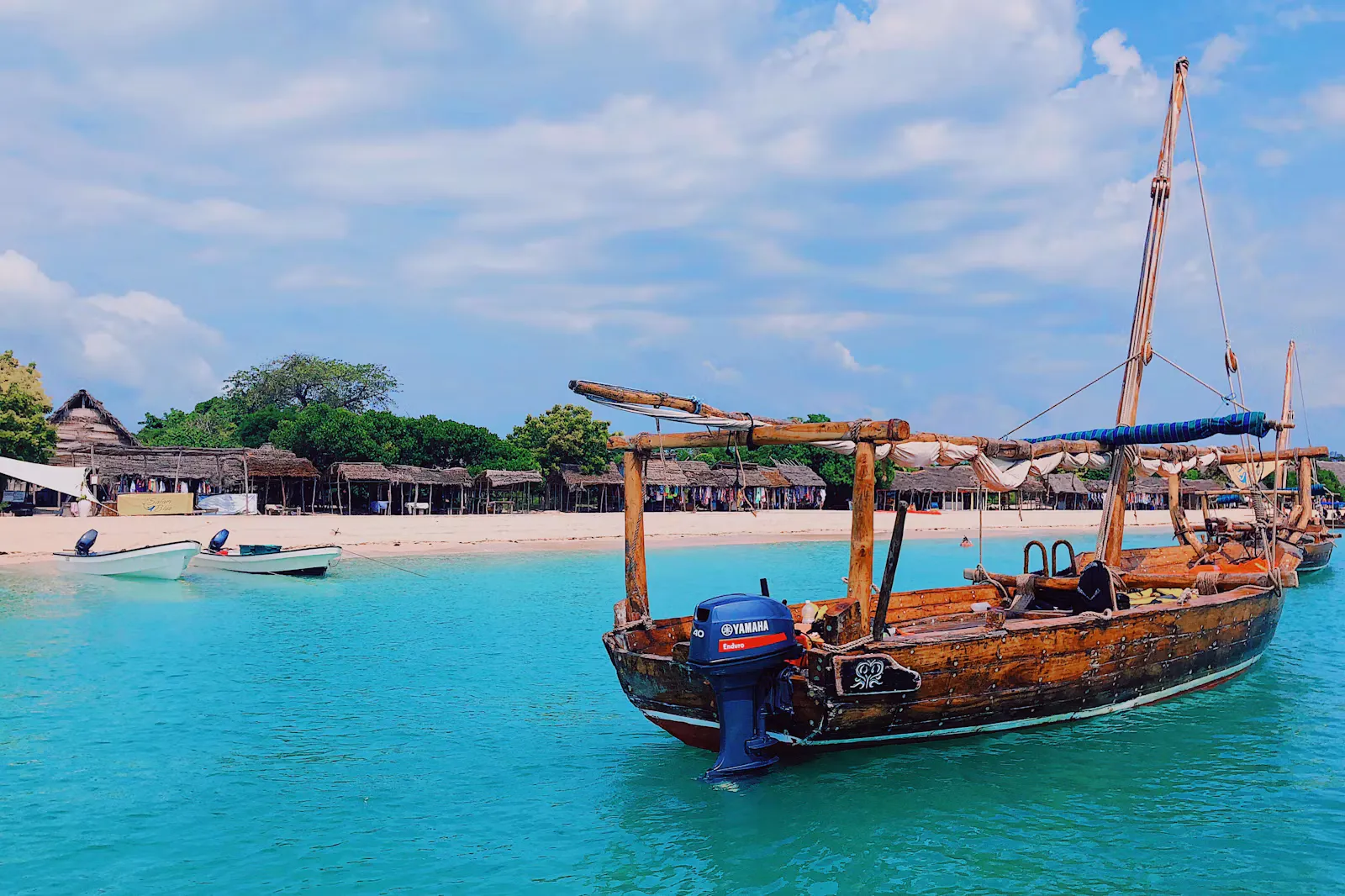 A wooden dhow with an outboard motor moored in turquoise water off a sandy beach with thatched huts under a blue sky