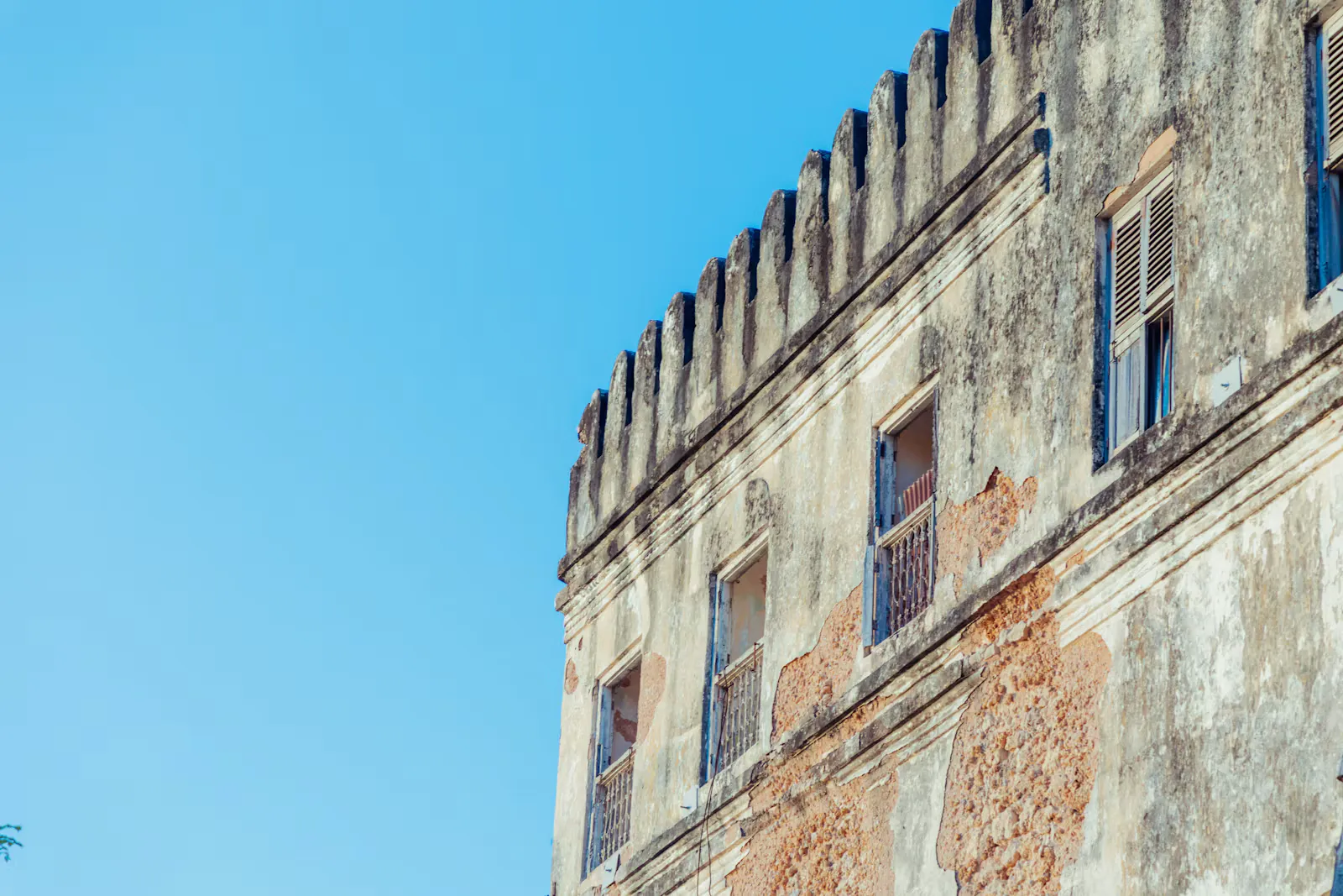 Verwitterte Fassade eines alten Gebäudes mit Zinnen und blauen Fensterläden unter blauem Himmel in Stone Town auf Sansibar