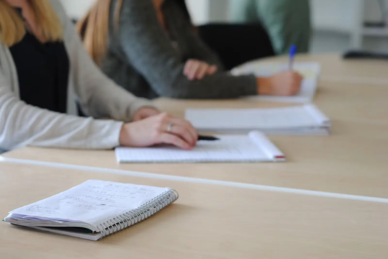Two students sit at a long light wood table in a bright room, writing with pens on spiral notepads and loose stapled sheets, with a closed notebook in the foreground
