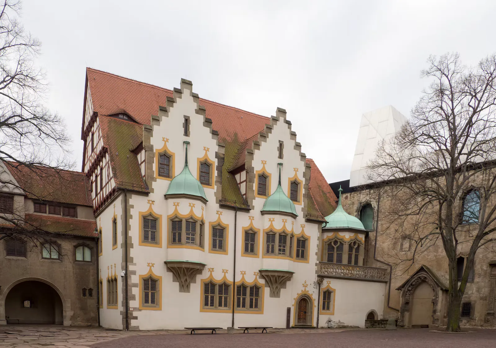 Weißes Gebäude mit Treppengiebeln, gelb umrandeten Fenstern und grünen Erkerhauben im Innenhof einer alten Burganlage, rechts ein moderner weißer Aufbau