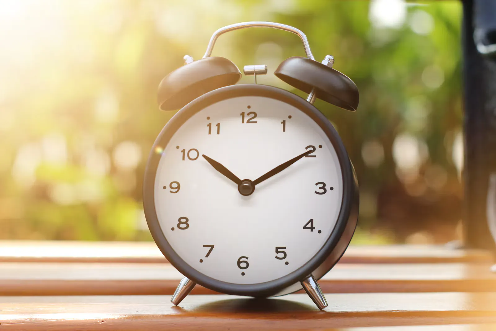 Schwarzer Wecker mit weissem Zifferblatt auf einer Holzbank im Morgenlicht, Symbol für den Jetlag und die verschobene innere Uhr