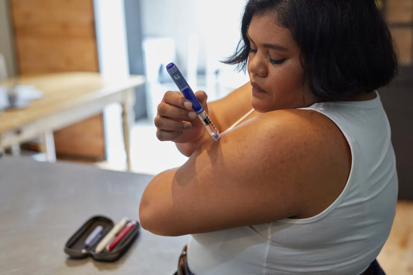 A woman in a white top injecting insulin into her upper arm with an insulin pen, with an open pen case on the table beside her
