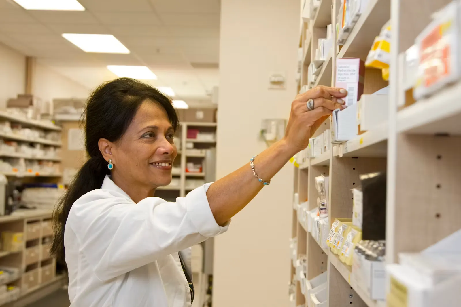 A doctor in a white coat reaching up for a boxed injectable medicine on a hospital pharmacy shelf