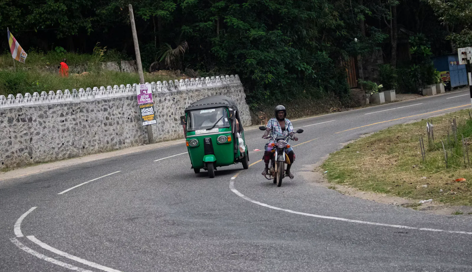 Grünes Tuk-Tuk und ein Motorradfahrer mit Helm in einer Kurve auf einer Landstraße mit Linksverkehr, daneben eine weiße Mauer und tropischer Wald