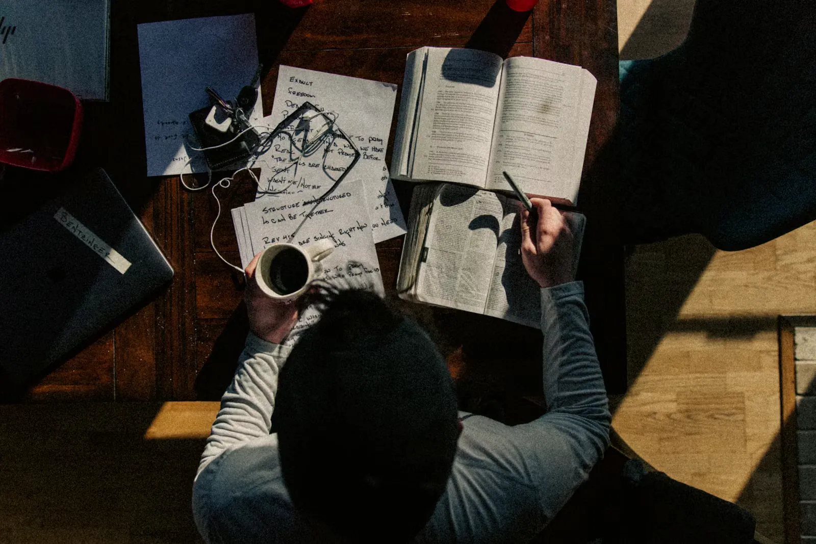 Blick von oben auf einen Schreibtisch, an dem jemand mit Kaffeetasse in der Hand Notizen in ein aufgeschlagenes Buch schreibt