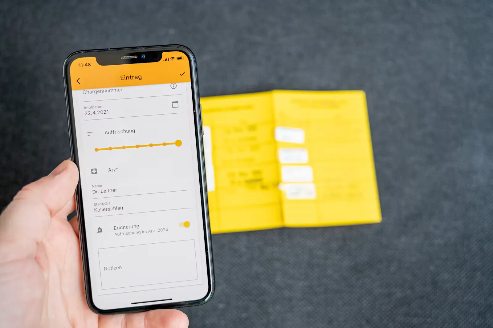 Hand hält ein Smartphone mit einer deutschsprachigen Impf-App, im Hintergrund liegt unscharf ein aufgeschlagener gelber Impfpass auf grauem Untergrund