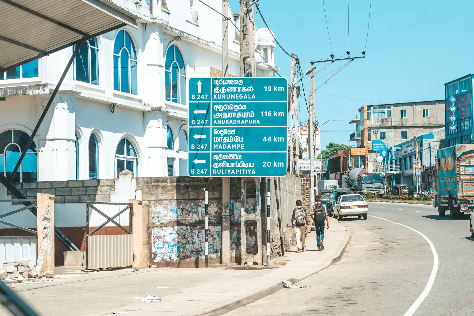 A blue road sign in Sri Lanka listing towns in Sinhala, Tamil and English, with two backpackers walking along the pavement beside it