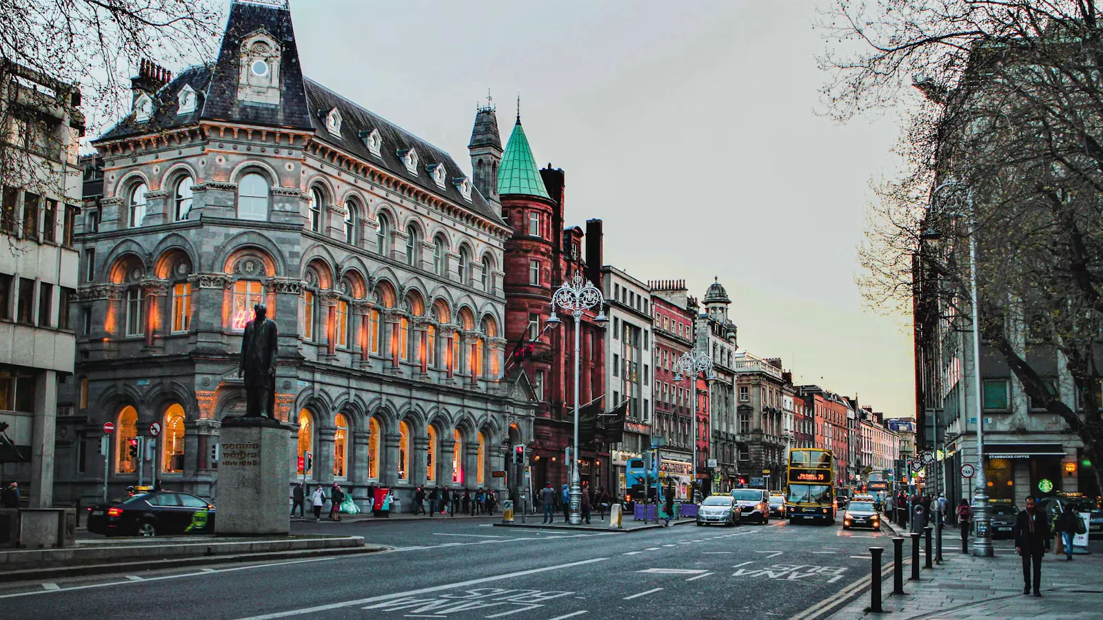 A wide Irish city street with a stone clock tower standing in the middle of the road
