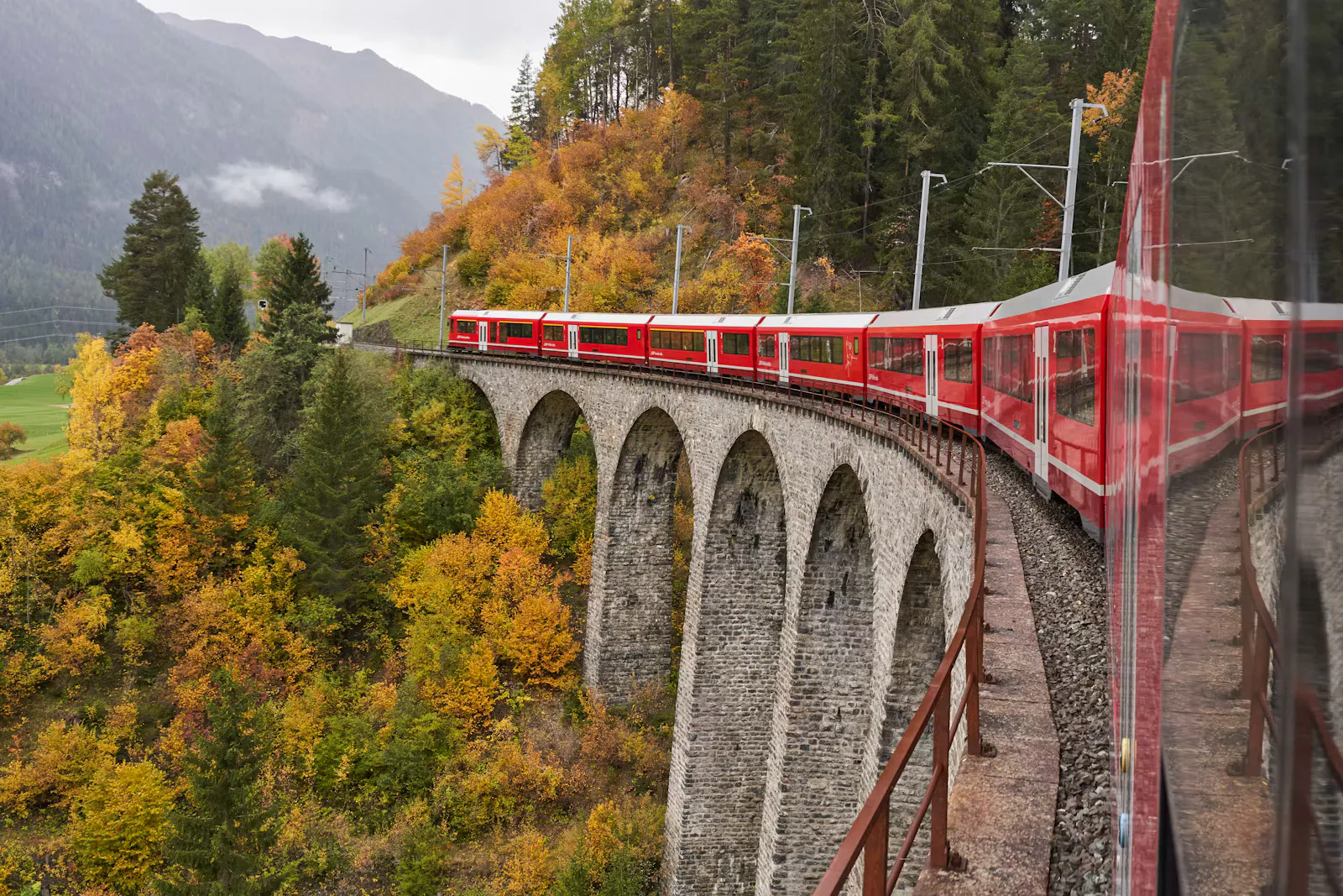 Roter Zug der Rhätischen Bahn auf einem steinernen Viadukt zwischen herbstlich gefärbten Bäumen, Symbolbild für die letzte Zugfahrt vor dem Auslandspraktikum