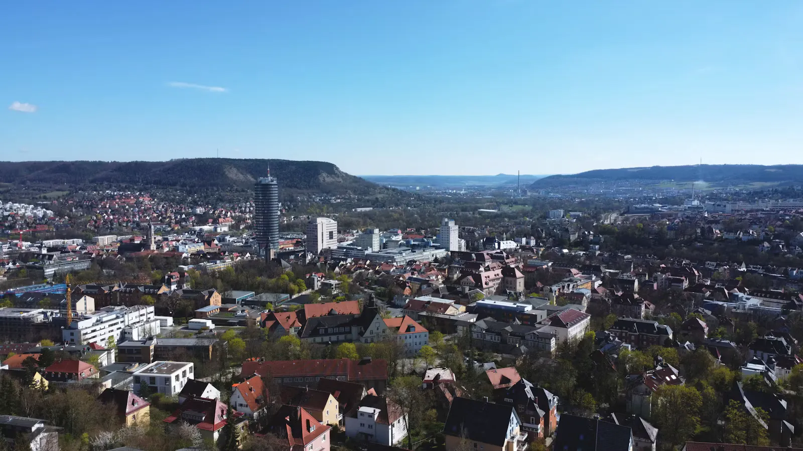 Luftbild von Jena mit dem runden JenTower in der Stadtmitte, roten Ziegeldächern und bewaldeten Bergrücken unter blauem Himmel