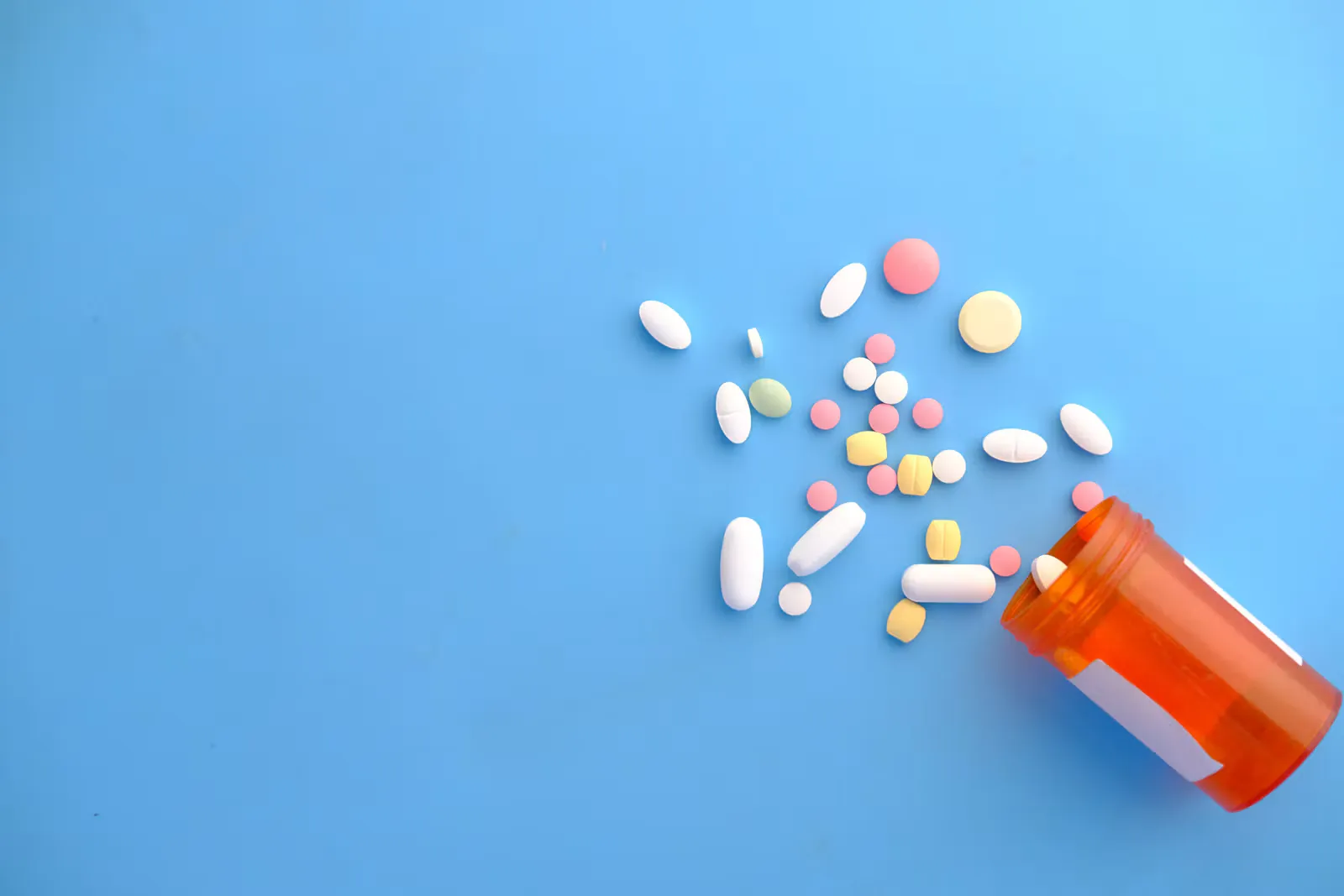 White, pink and yellow tablets and capsules spilling out of an orange pill bottle onto a light blue surface