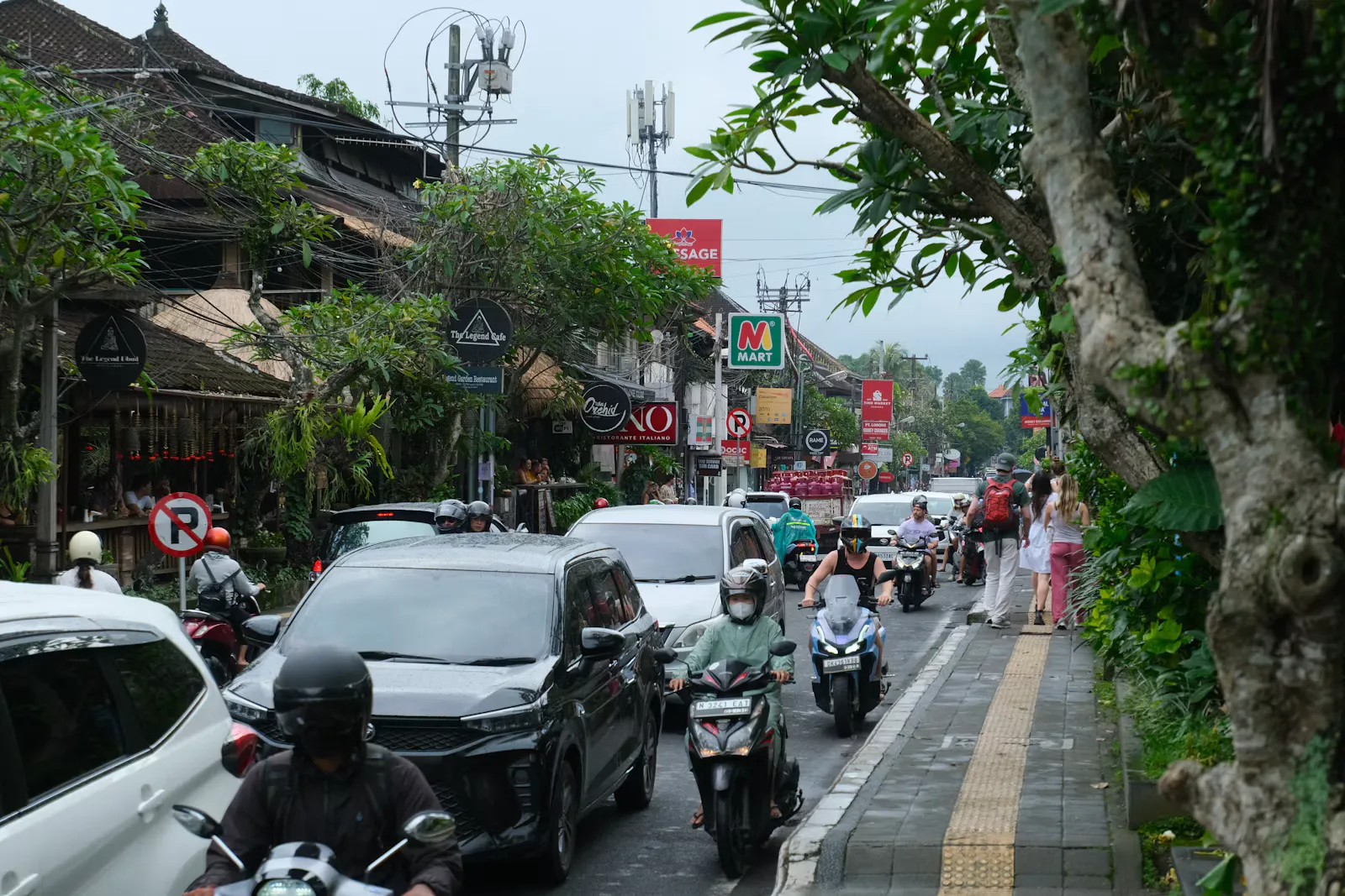 Dichter Verkehr aus Autos und Motorrollern auf einer Hauptstraße in Ubud auf Bali, Fußgänger gehen auf dem Gehweg daneben