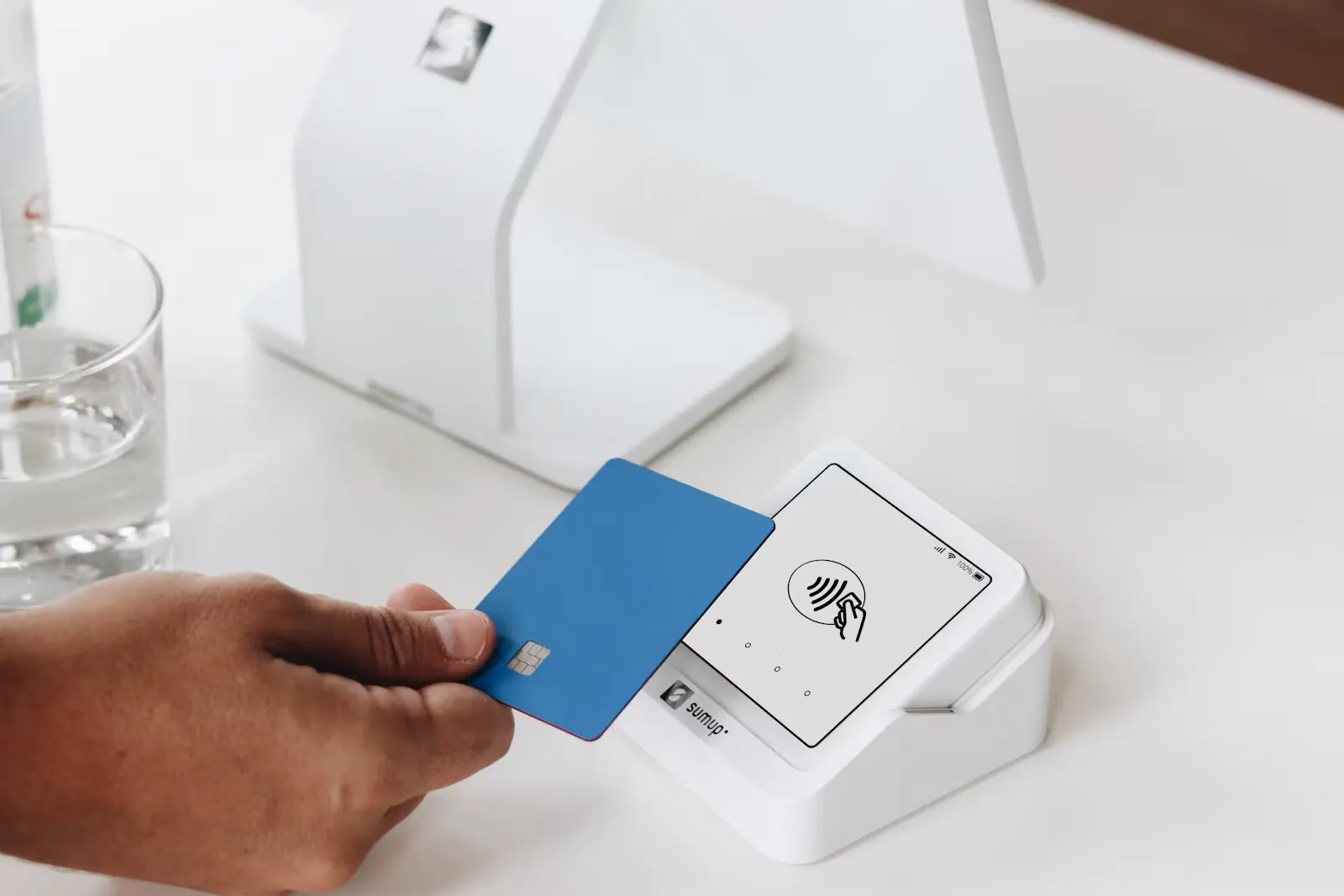 A hand holding a blue bank card next to a white card terminal on a desk, the kind of card payment that adds a gateway charge to NEET exam fees