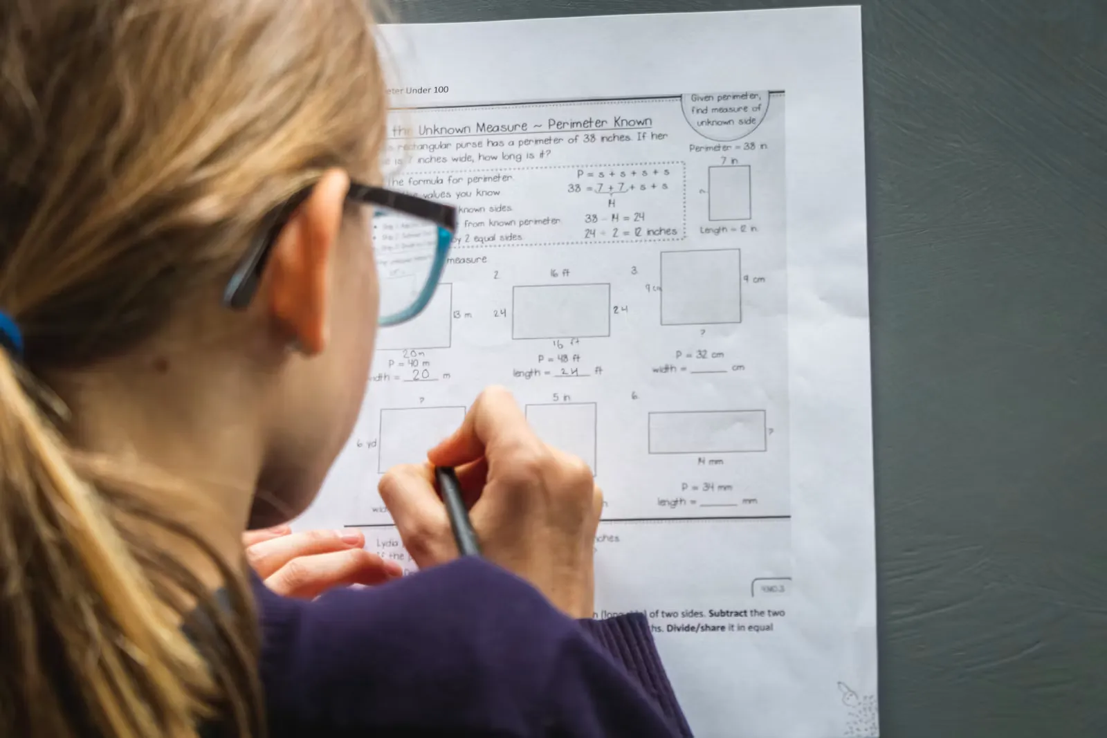 A school student in glasses working through a printed worksheet of problems, pen in hand