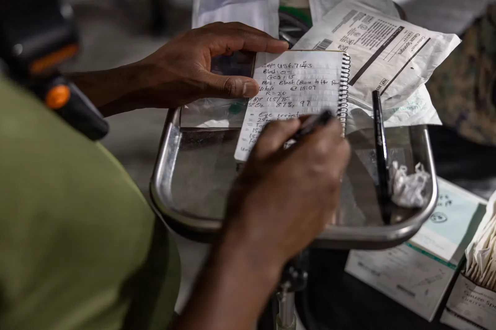 Hands writing clinical notes in a small spiral notebook resting on a metal tray next to medical supplies