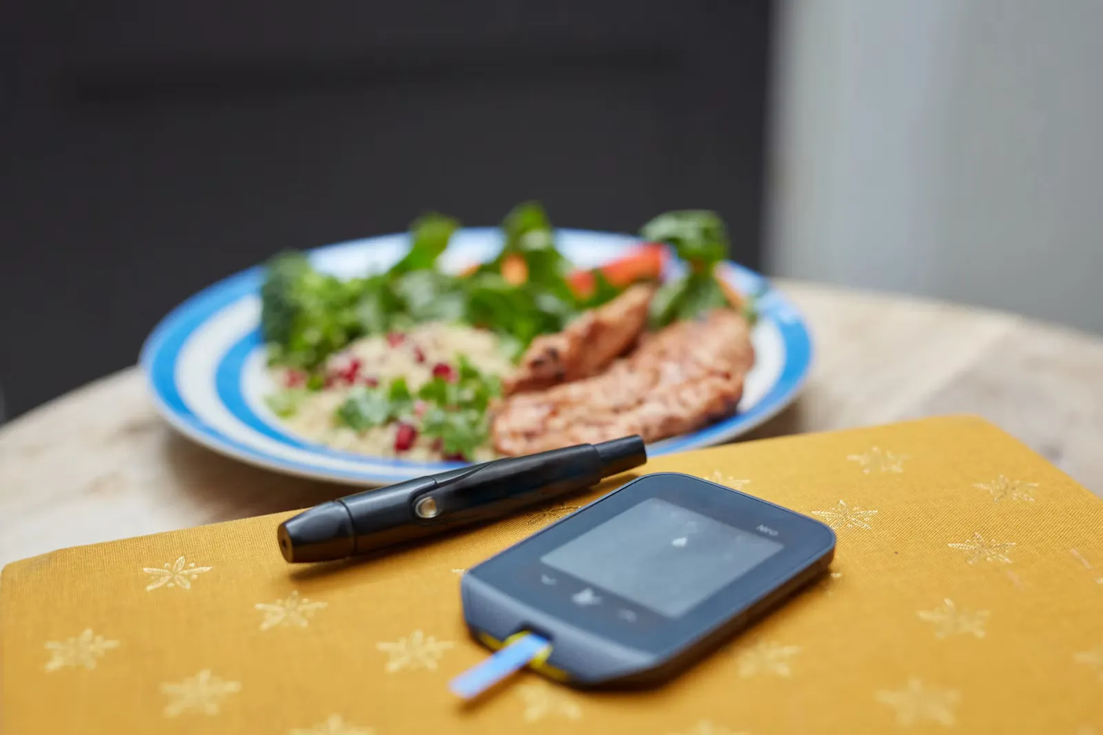 A blood glucose meter and a lancing device resting on a yellow notebook, with a plate of salad, rice and grilled chicken behind them