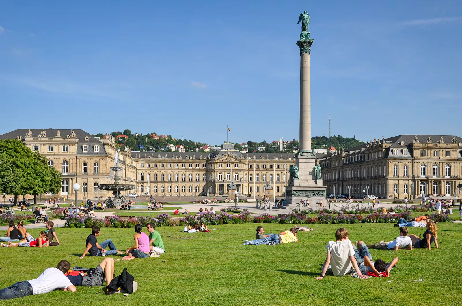 Studierende sitzen und liegen bei Sonnenschein auf dem Rasen des Stuttgarter Schlossplatzes, im Hintergrund das Neue Schloss und die Jubiläumssäule