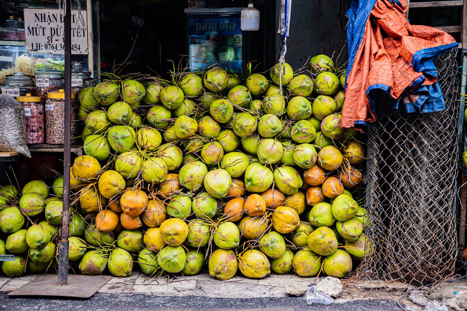 A large pile of green and orange coconuts stacked on the pavement in front of a small street market stall