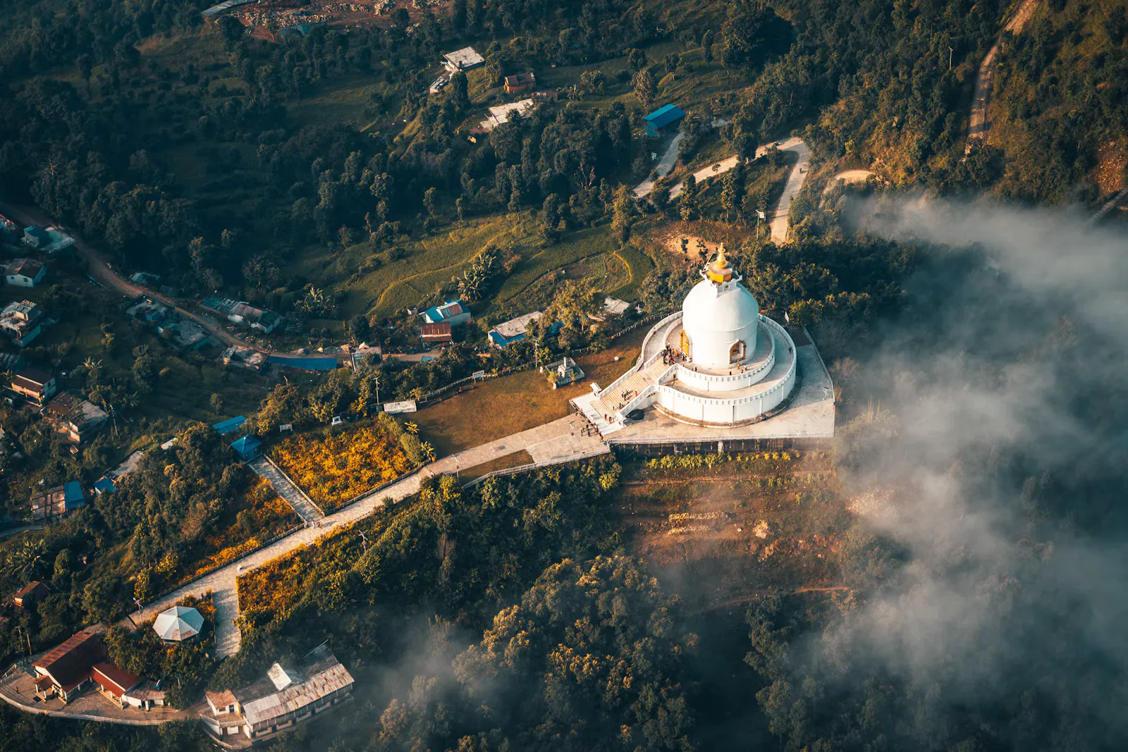 Luftaufnahme einer weißen Stupa auf einem bewaldeten Hügel bei Pokhara in Nepal, mit Häusern, Feldern und aufsteigendem Nebel