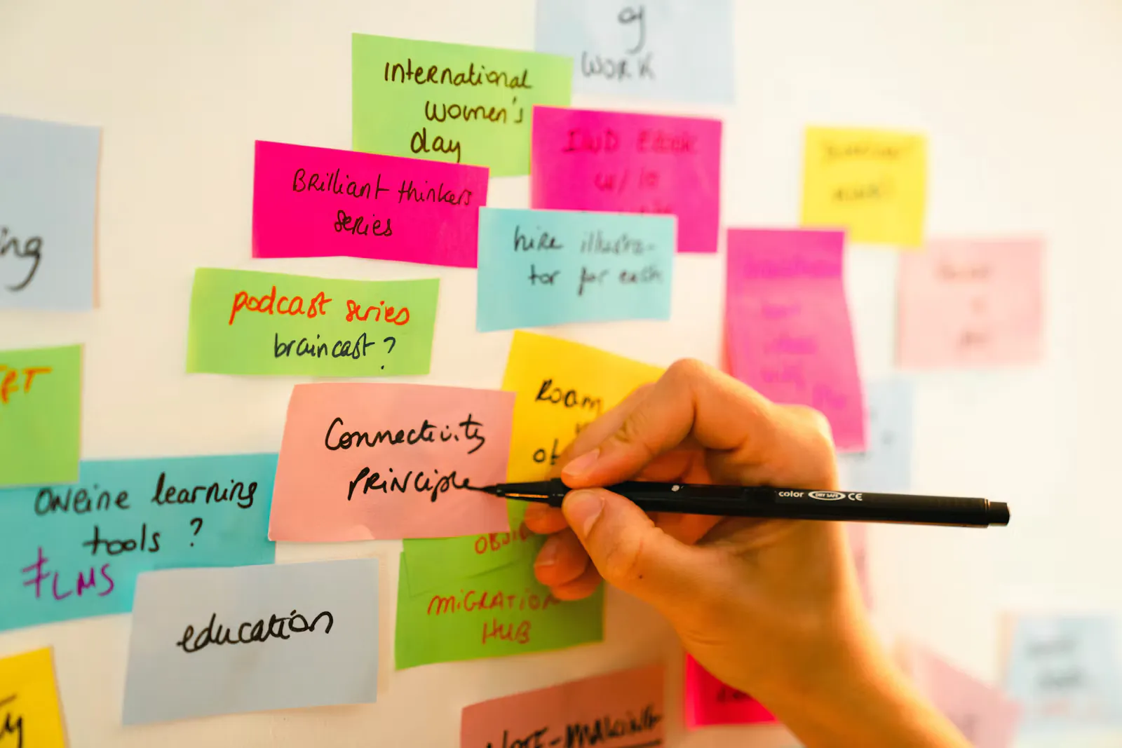 Hand schreibt mit schwarzem Stift auf eine rosa Haftnotiz an einer Wand voller bunter Notizzettel