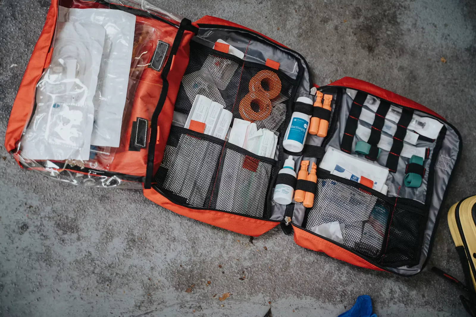 Geöffnete orange Erste-Hilfe-Tasche auf grauem Betonboden mit Verbandsmaterial, Kompressen und kleinen Fläschchen in Netzfächern