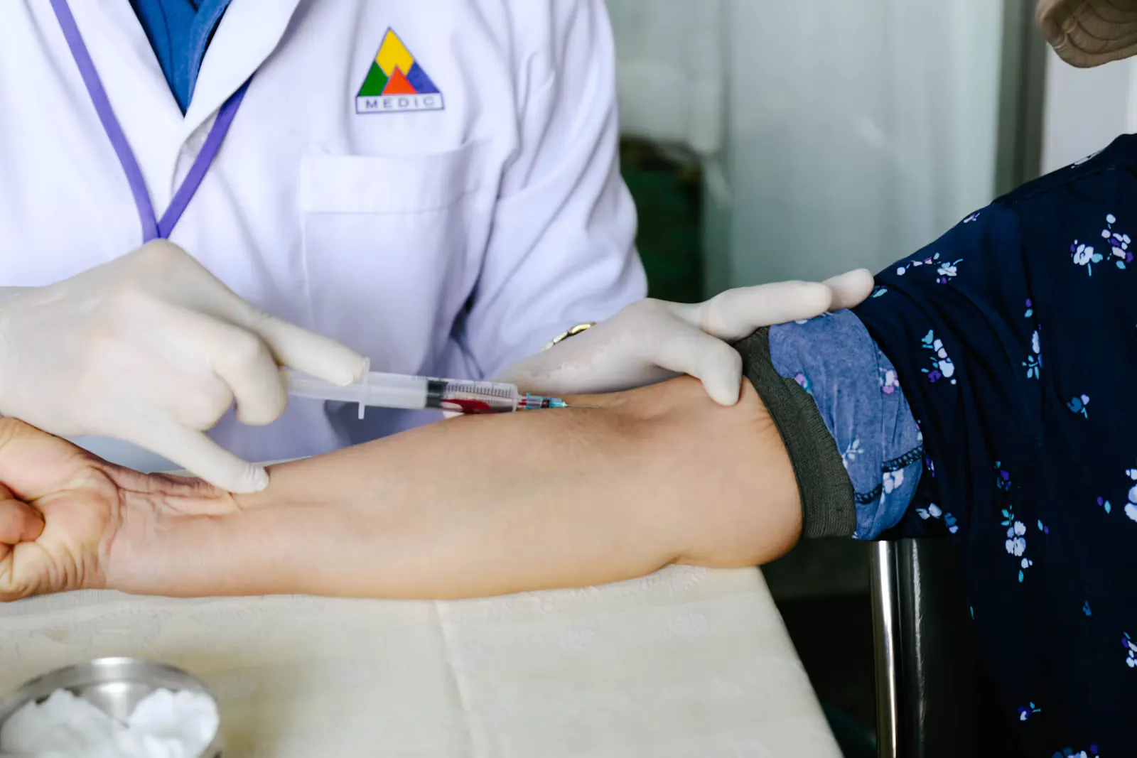 Eine Fachperson mit Einmalhandschuhen entnimmt mit einer Spritze Blut aus der Armvene einer sitzenden Person, deren Arm auf einem Tisch liegt, Beispiel für venöse Blutentnahme im Klinikalltag