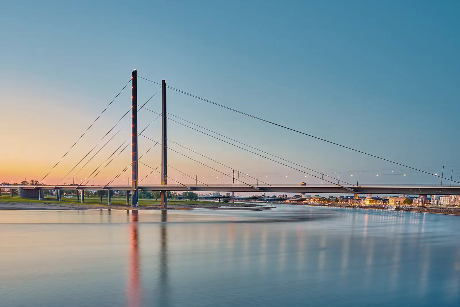 Schrägseilbrücke über den Rhein in Düsseldorf im Abendlicht, rechts die beleuchtete Uferpromenade der Altstadt, das ruhige Wasser spiegelt die Lichter