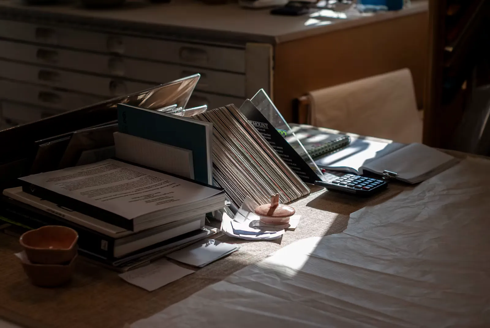 Sonnenlicht fällt auf einen Holztisch mit gestapelten Büchern, Mappen und Unterlagen in einem Ablagefach, rechts liegt ein Taschenrechner auf einem Notizbuch