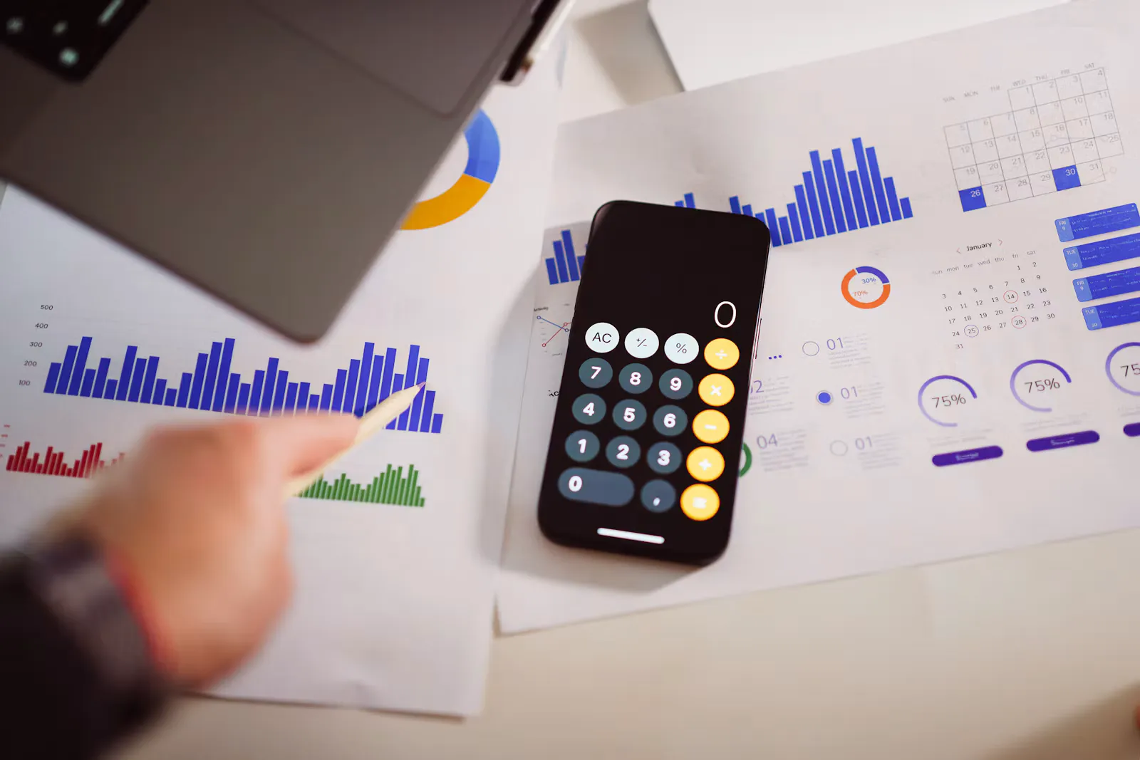 Hand mit Stift zeigt auf Diagramme, daneben ein Smartphone mit geöffnetem Taschenrechner auf ausgedruckten Auswertungen