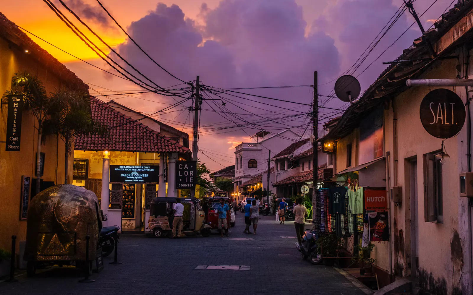 Abendstimmung in einer Gasse der Altstadt von Galle mit Tuk-Tuks, Cafés, Stromleitungen und violettem Himmel