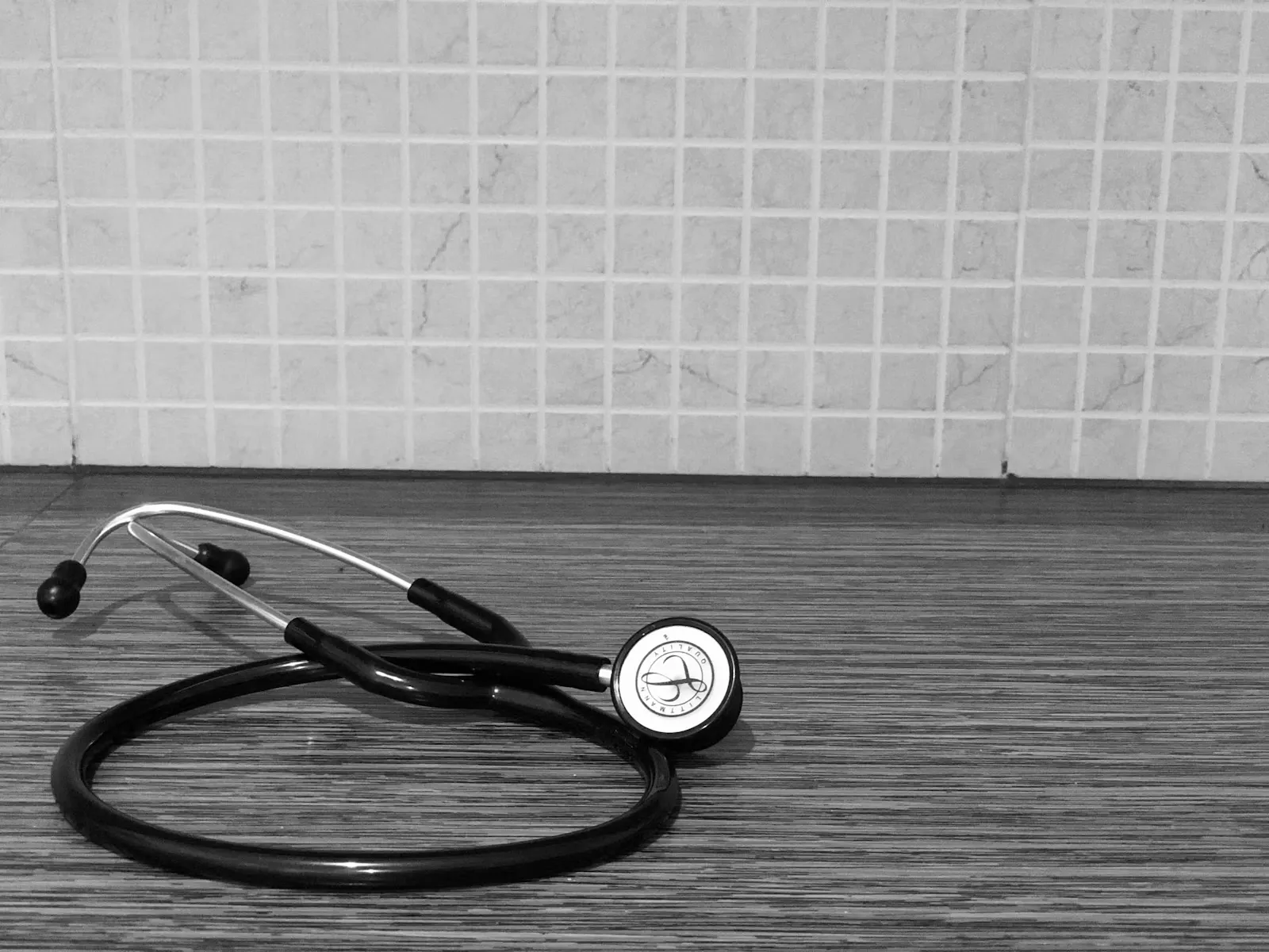 A black and white photo of a stethoscope lying on a wooden counter in front of a tiled wall
