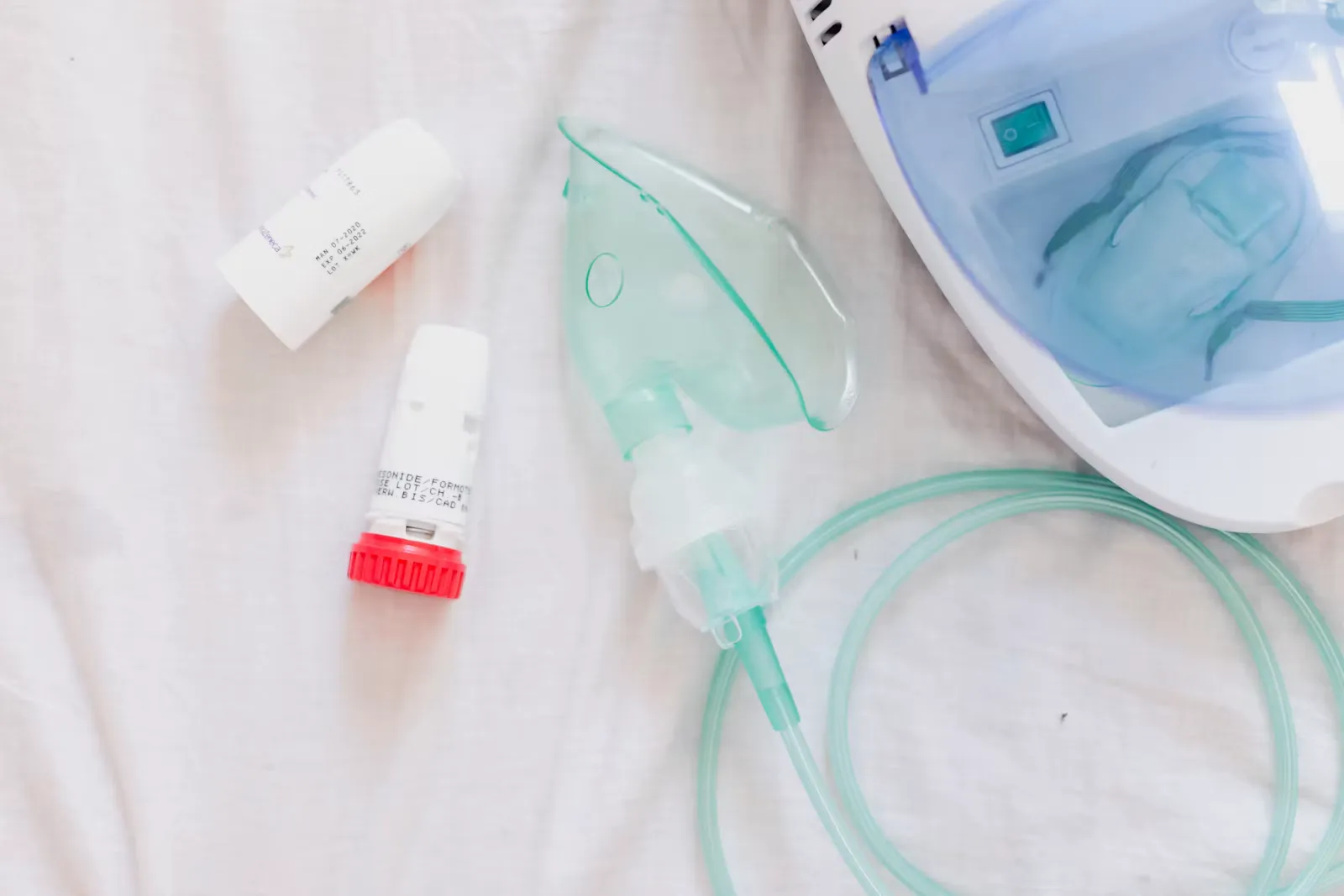 A nebuliser machine with a green child's face mask and tubing next to two inhalers on a white sheet