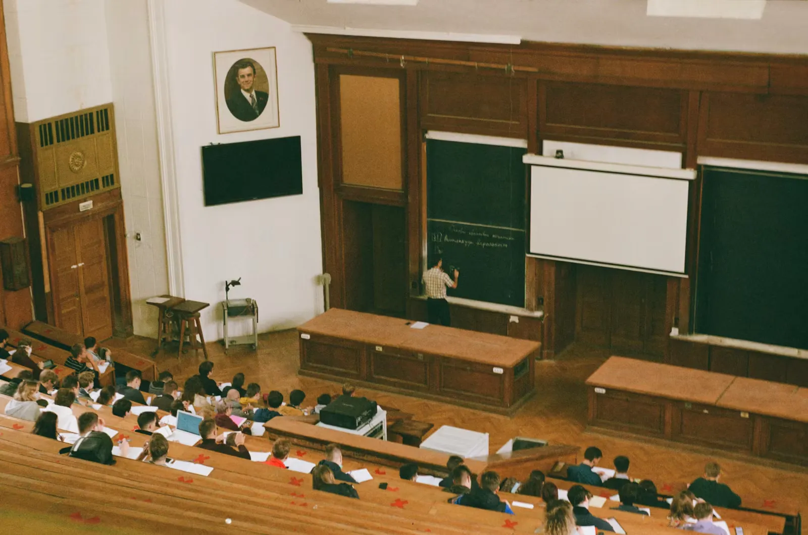 Students taking notes in a wood-panelled European university lecture hall while a lecturer writes on the blackboard