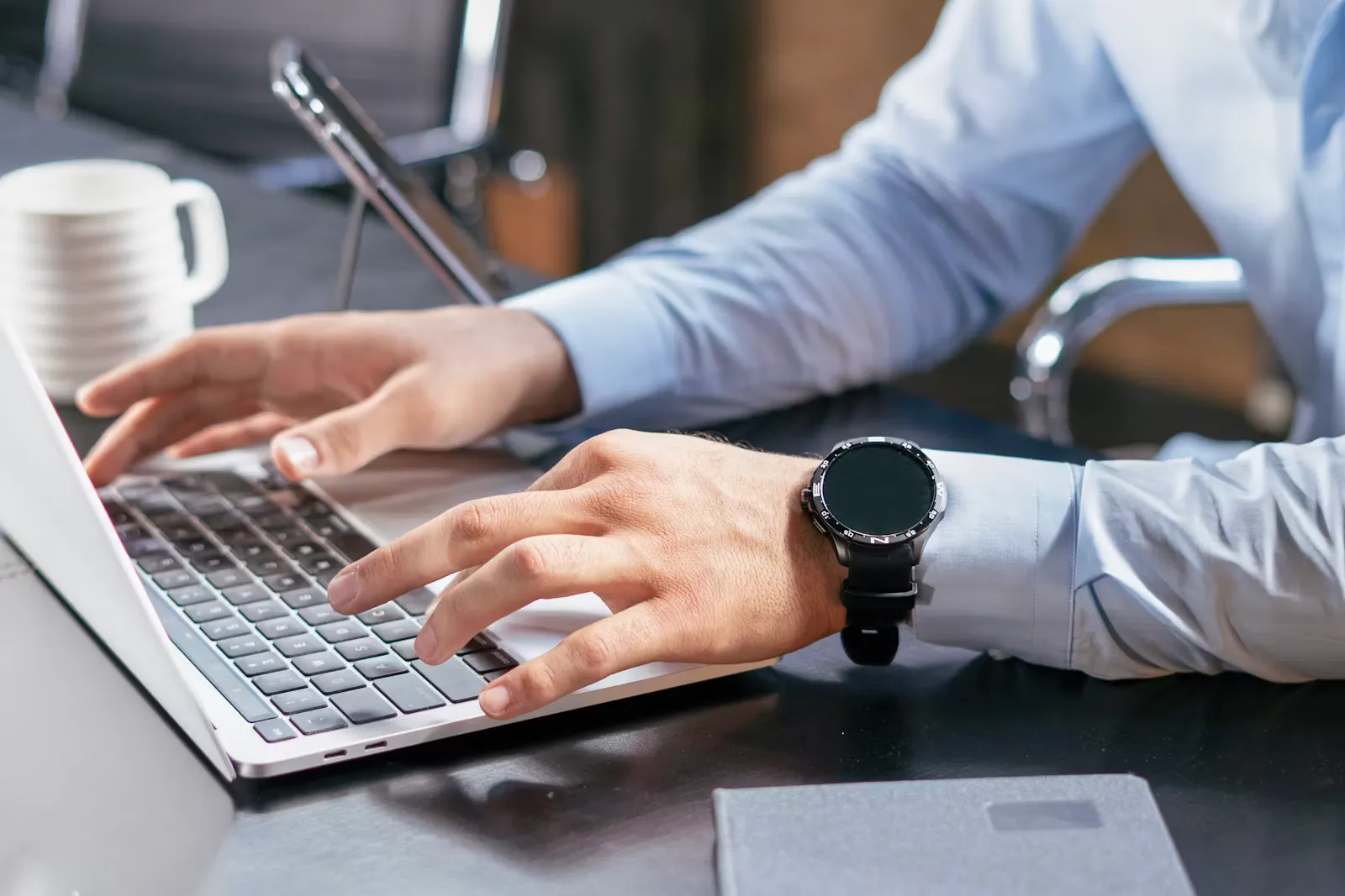 Close-up of a person's hands typing on a silver laptop keyboard at a dark desk, with a white mug and a closed notebook beside them