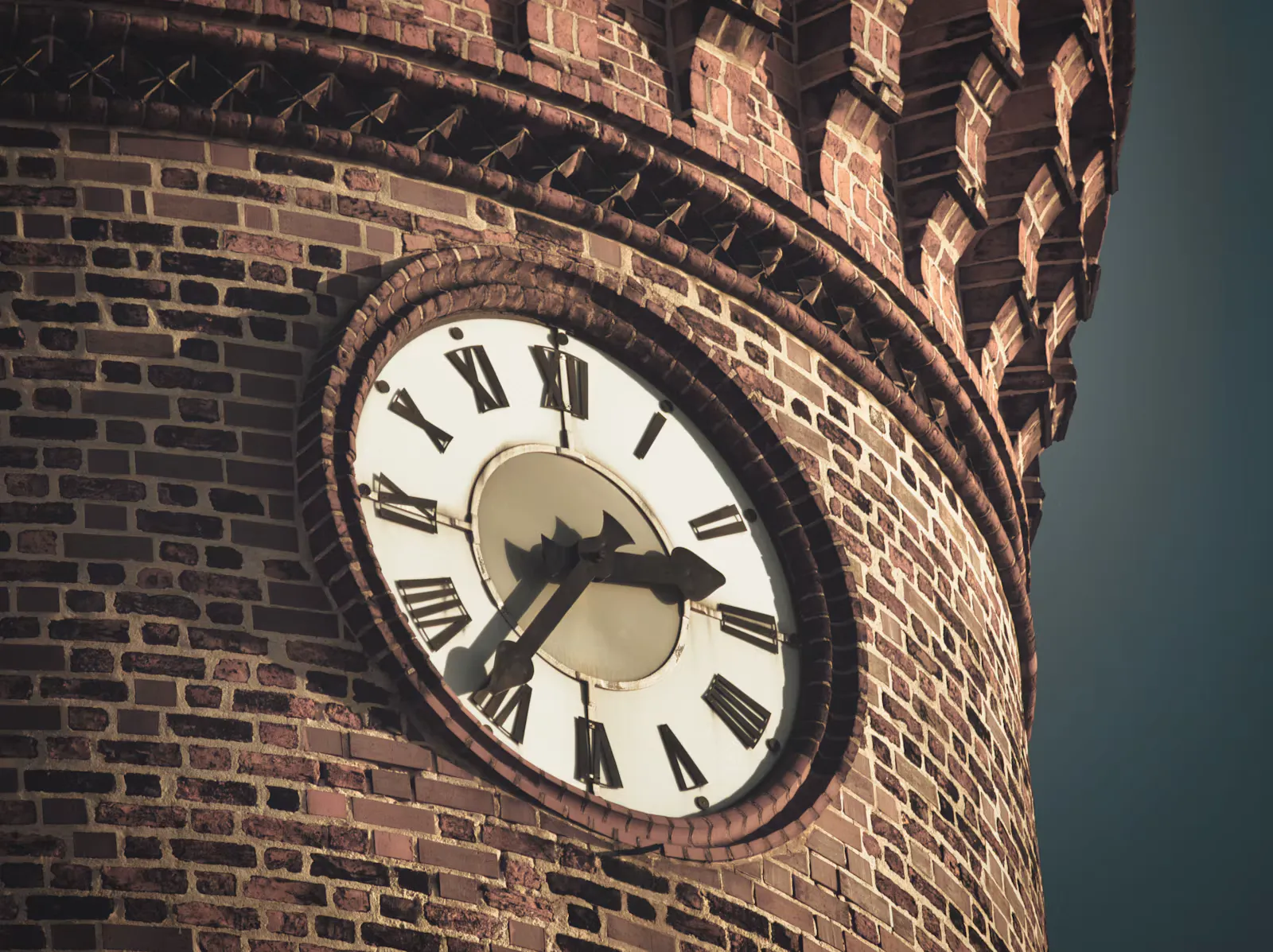 Runde Turmuhr mit römischen Ziffern an einem alten Backsteinturm mit Zinnenkranz vor dunklem Himmel