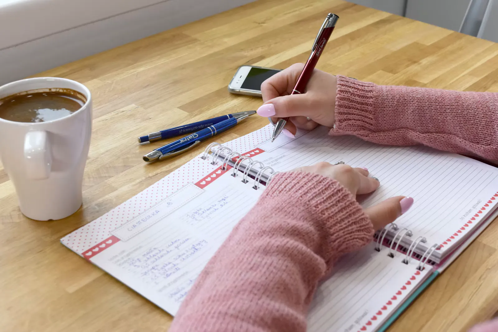 Hände einer Person in rosa Pullover, die neben einer Tasse Kaffee Notizen in einen Planer schreibt, Symbolbild für die Nachbereitung nach dem Kulturschock