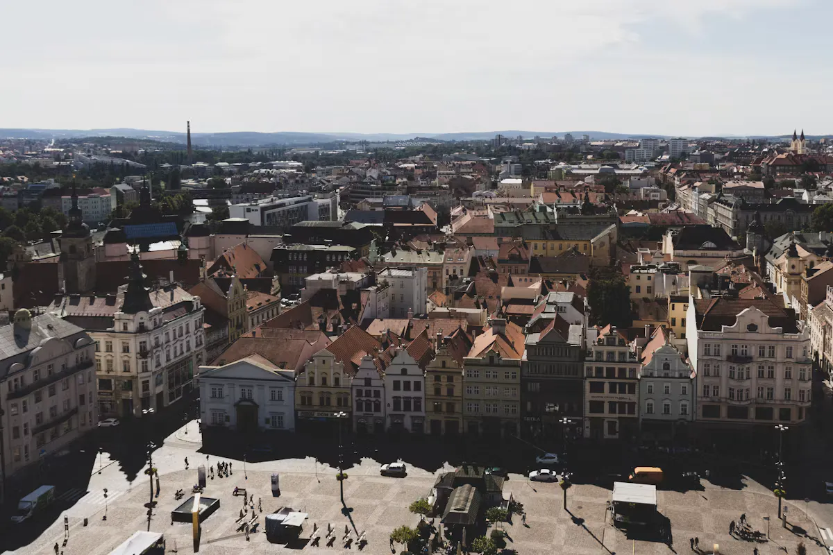 Pilsen's main square seen from above, with pastel historic townhouses, red tiled roofs and the city spreading towards green hills