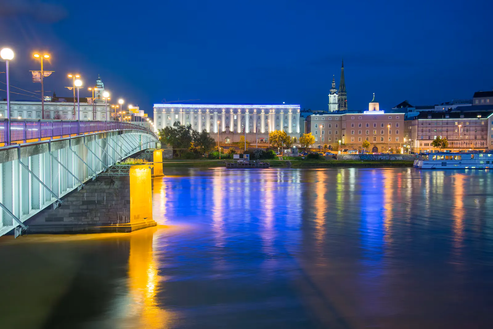 Nibelungenbrücke über die Donau in Linz bei Nacht mit beleuchteten Gebäuden und Kirchtürmen am Ufer, Symbolbild für die Planung der Famulatur an der JKU