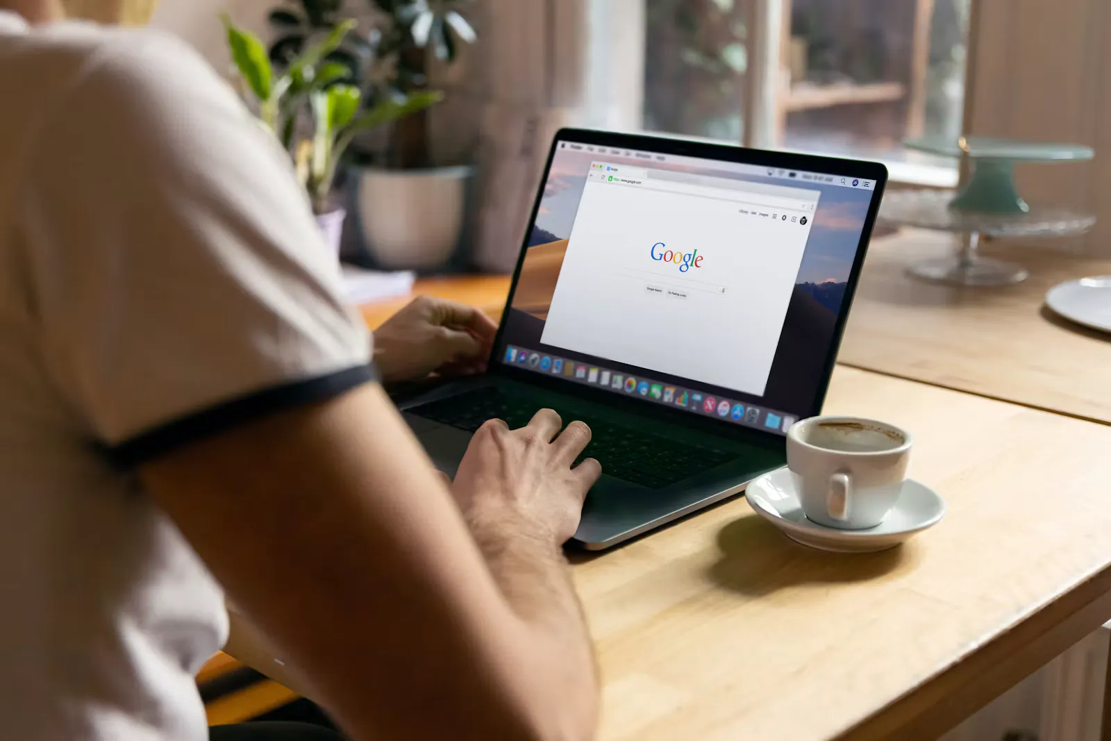 Person tippt an einem Laptop auf einem Holztisch, daneben eine Tasse Kaffee auf einer Untertasse
