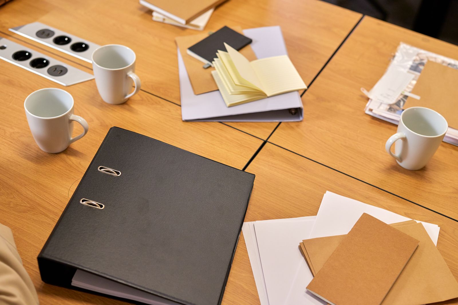 Schwarzer Ordner, Notizbücher, Papierstapel und zwei Kaffeetassen liegen auf einem hellen Holztisch bei der Vorbereitung der Praktikumsunterlagen