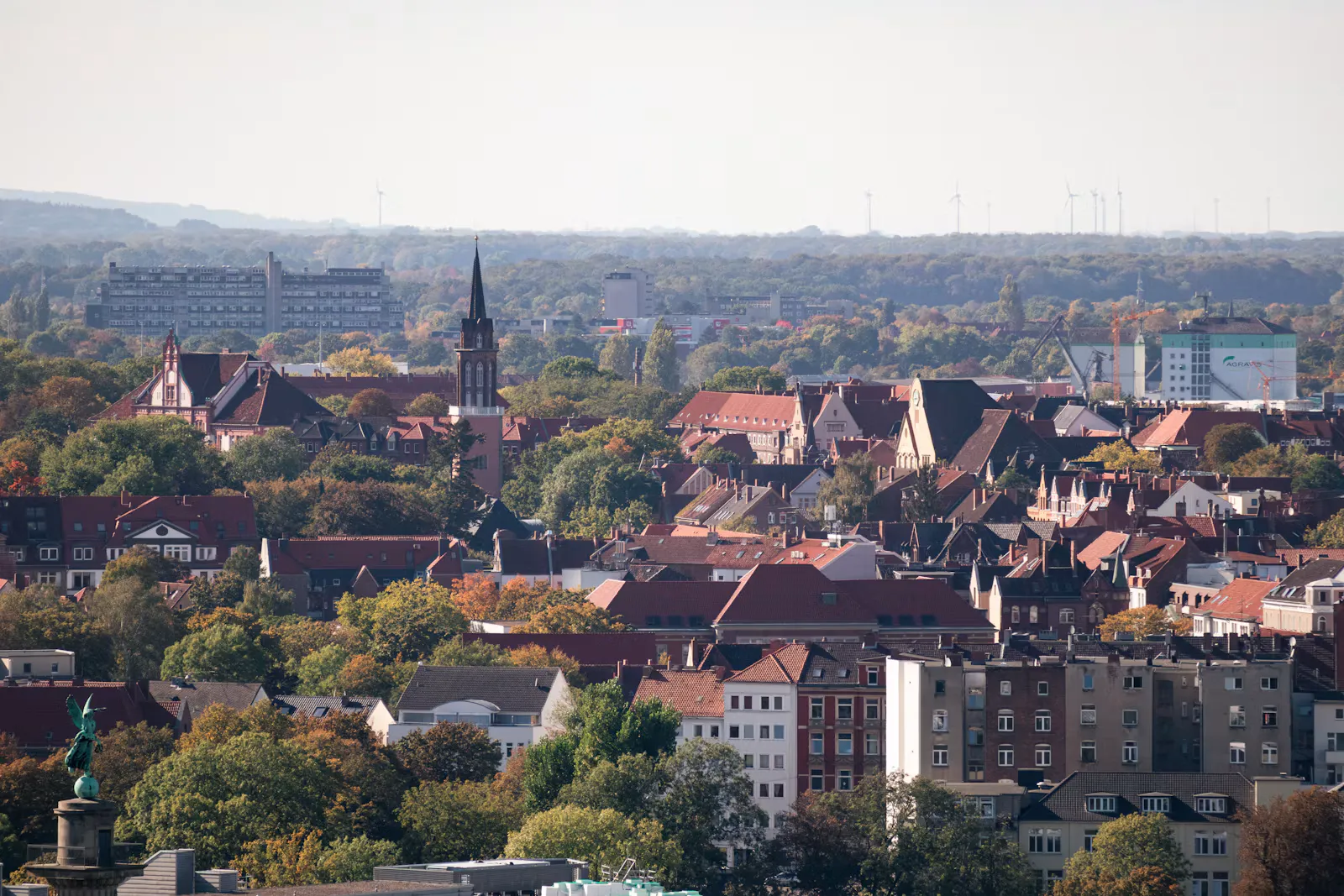 Blick über rote Ziegeldächer einer norddeutschen Stadt mit spitzem Kirchturm und herbstlich gefärbten Bäumen, im Hintergrund Wald und Windräder
