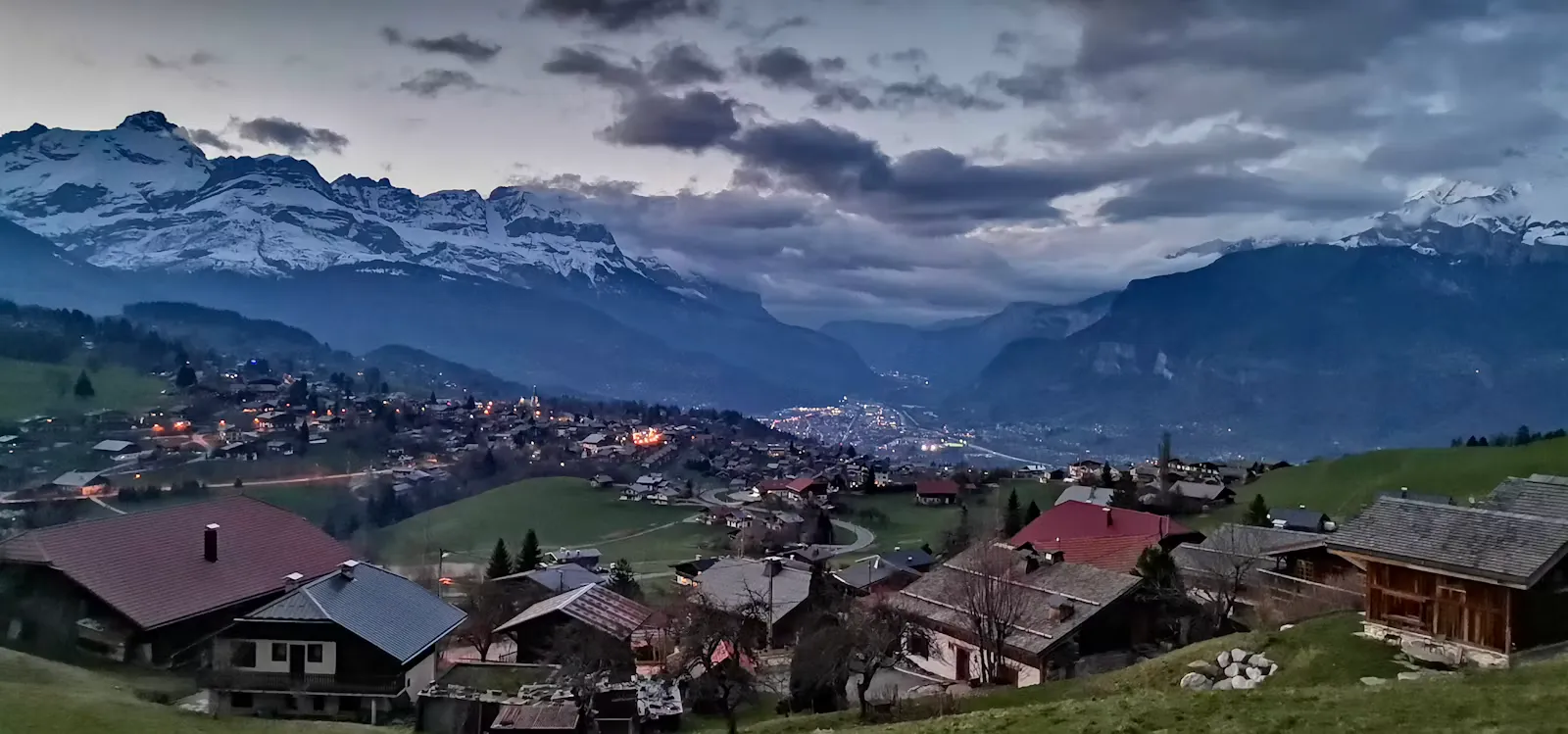 Dorf mit Holzhäusern an einem Berghang in der Abenddämmerung, im Tal leuchten die Lichter einer Stadt unter verschneiten Gipfeln