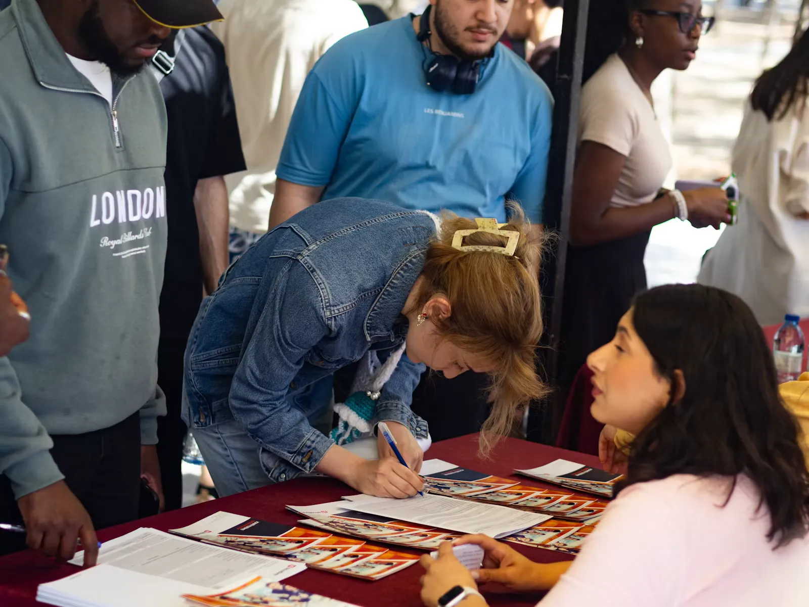 Young woman leaning over a table to fill in a form while others wait in a queue behind her