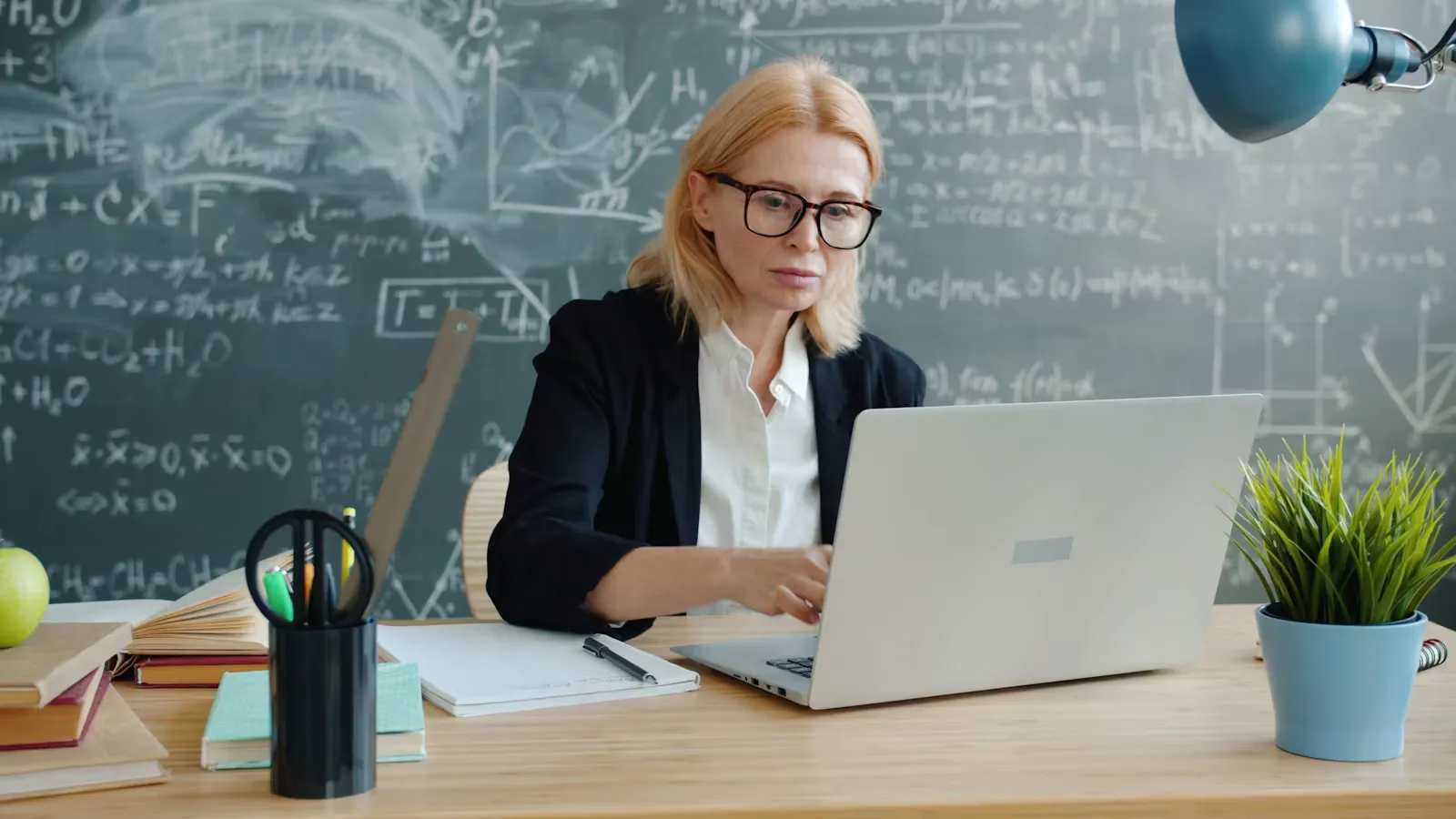 Doctor sitting at a desk typing on a laptop in front of a large chalkboard, working through an online exam application
