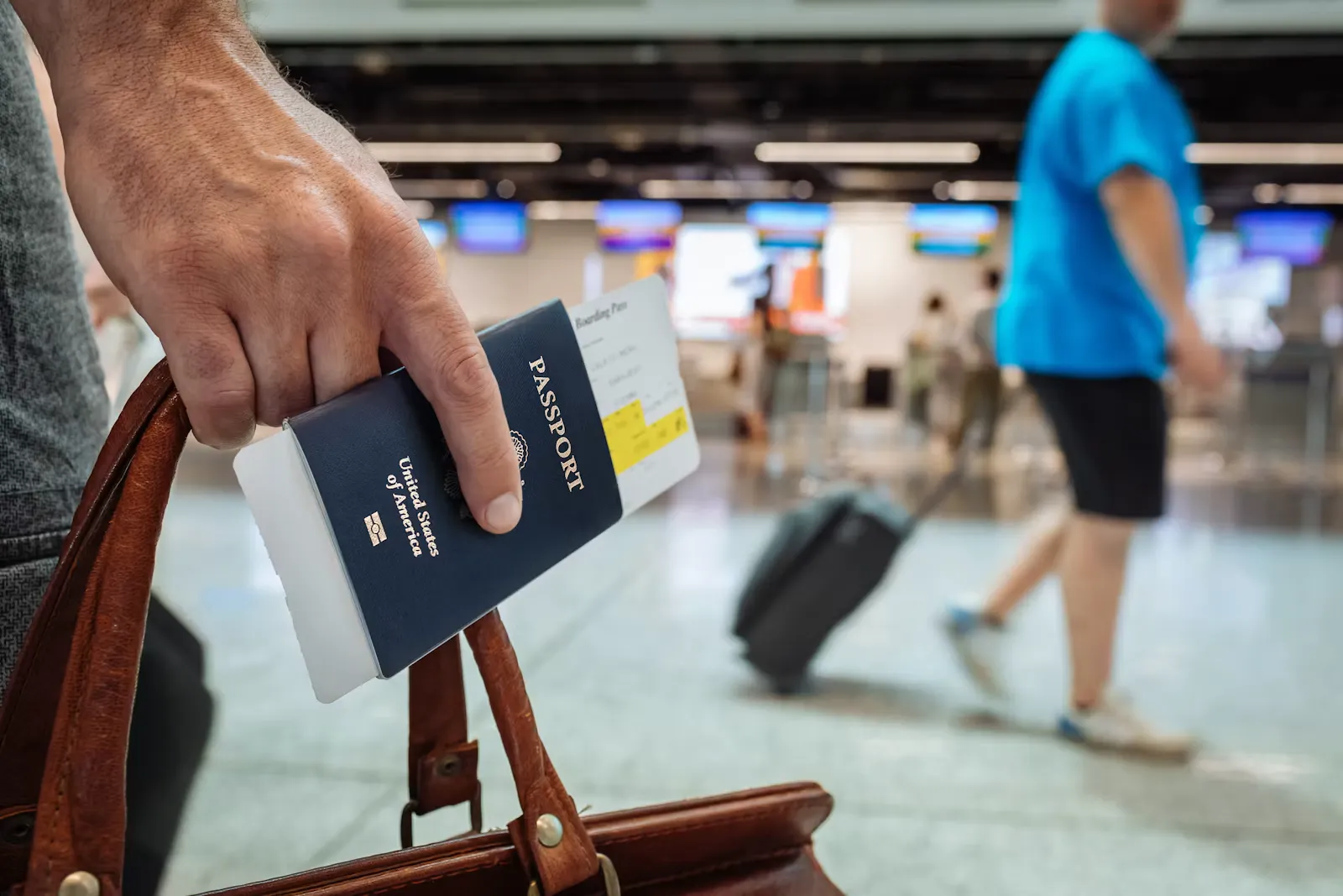 Hand mit Reisepass und Bordkarte über einer Ledertasche vor Abflugschaltern