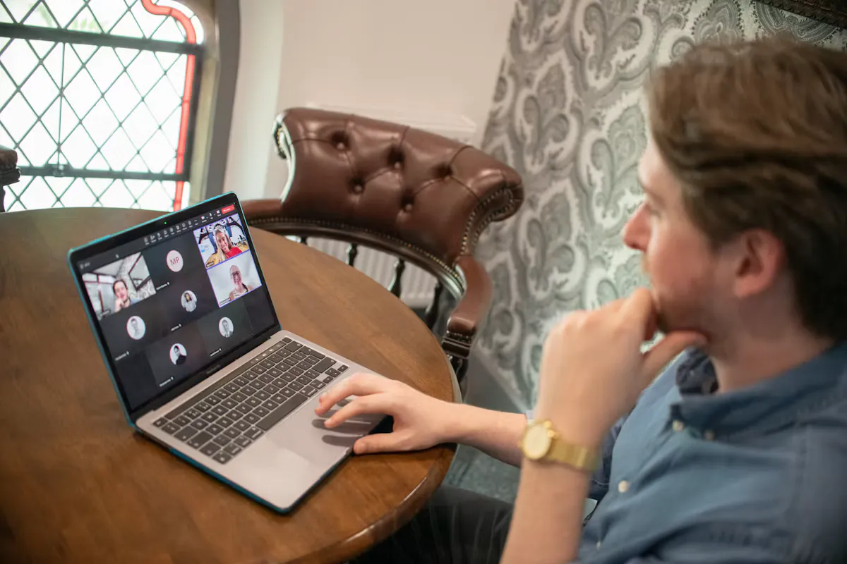 Young man at a wooden table looking at a laptop with a video call grid on screen, like an online admission interview