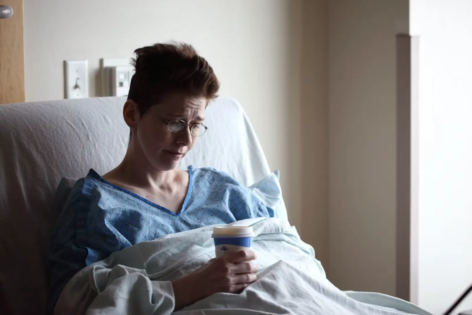 Junge Frau sitzt auf einem Bett und trinkt aus einem Becher, neben ihr eine zerknuellte Decke