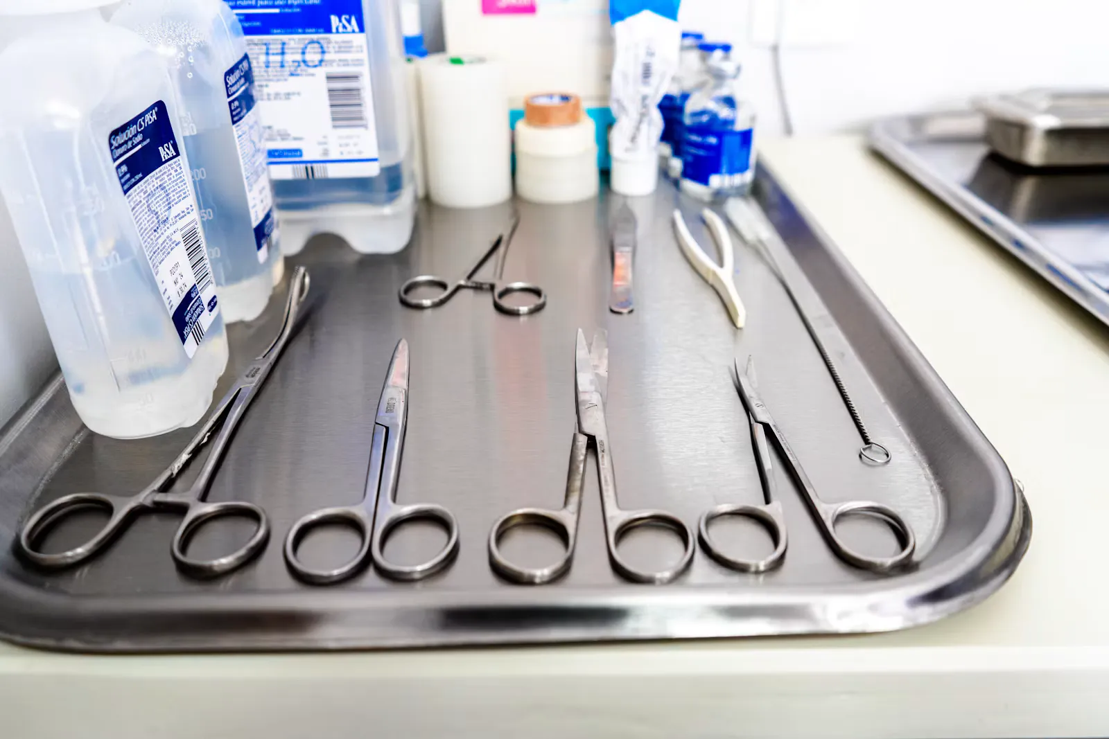 A stainless steel tray with surgical scissors, artery forceps and a needle holder laid out next to bottles of sterile water