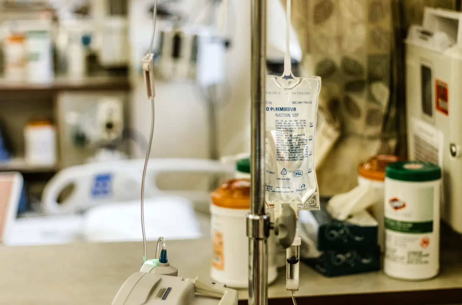An IV fluid bag hanging on a drip stand at a hospital bedside, with medicine bottles and equipment on the shelf behind it