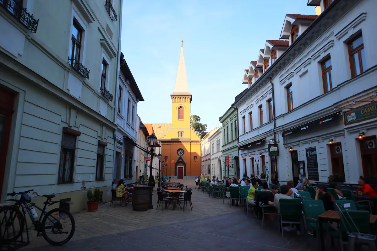 An old town street in Košice in the evening sun, with outdoor cafe tables, pastel houses and a yellow church tower at the end of the street